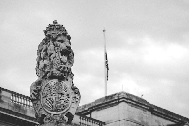 A stone lion wearing a crown and a coat of arms stands in front of a flag and a parliament building